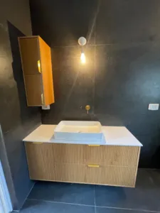 ארון אמבטיה בצבע עץ בהיר וידיות בצבע זהב עם קיר בצבע שחור
