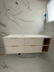 ארון אמבטיה בצבע ורוד בהיר עם ידיות זהב חלון וקיר בצבע לבן עם נגיעות של אפור